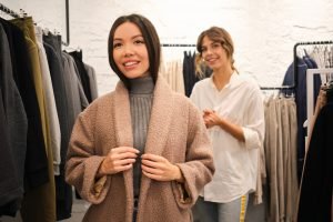 Beautiful stylish Asian girl joyfully trying on new casual coat in clothes store