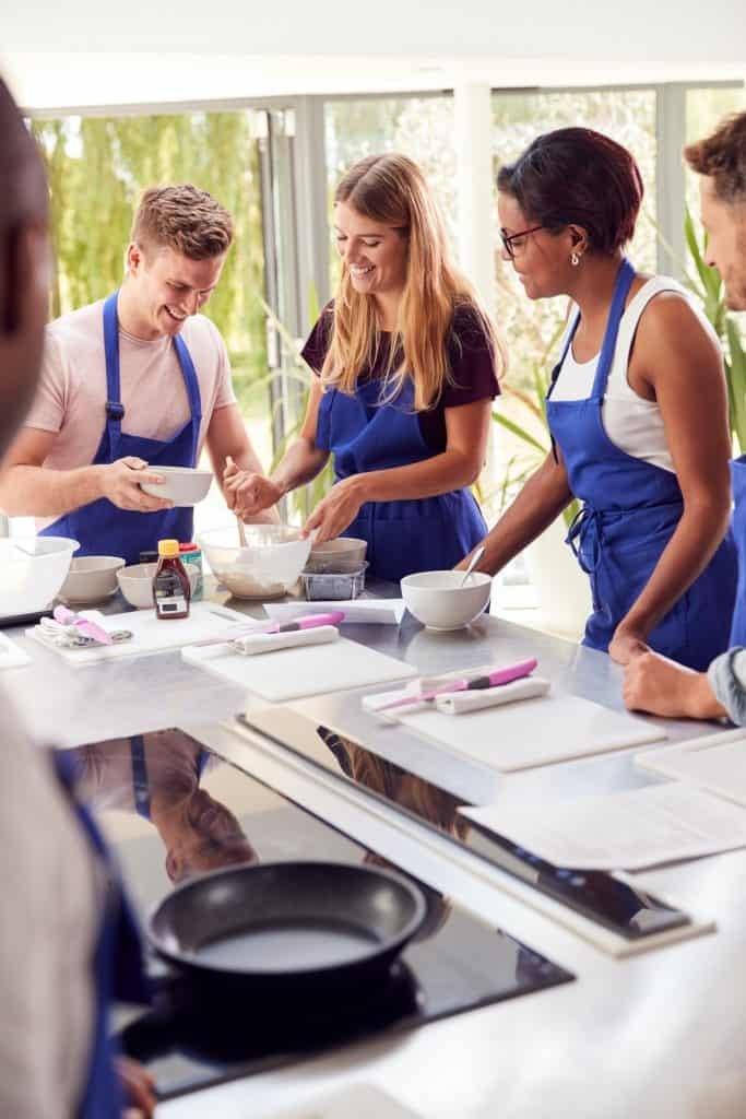 Female Teacher With Male Student Mixing Ingredients For Recipe In Cookery Class In Kitchen