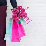 Stylish man holding flowers and pink, blue shopping bags on brick background. Woman's day