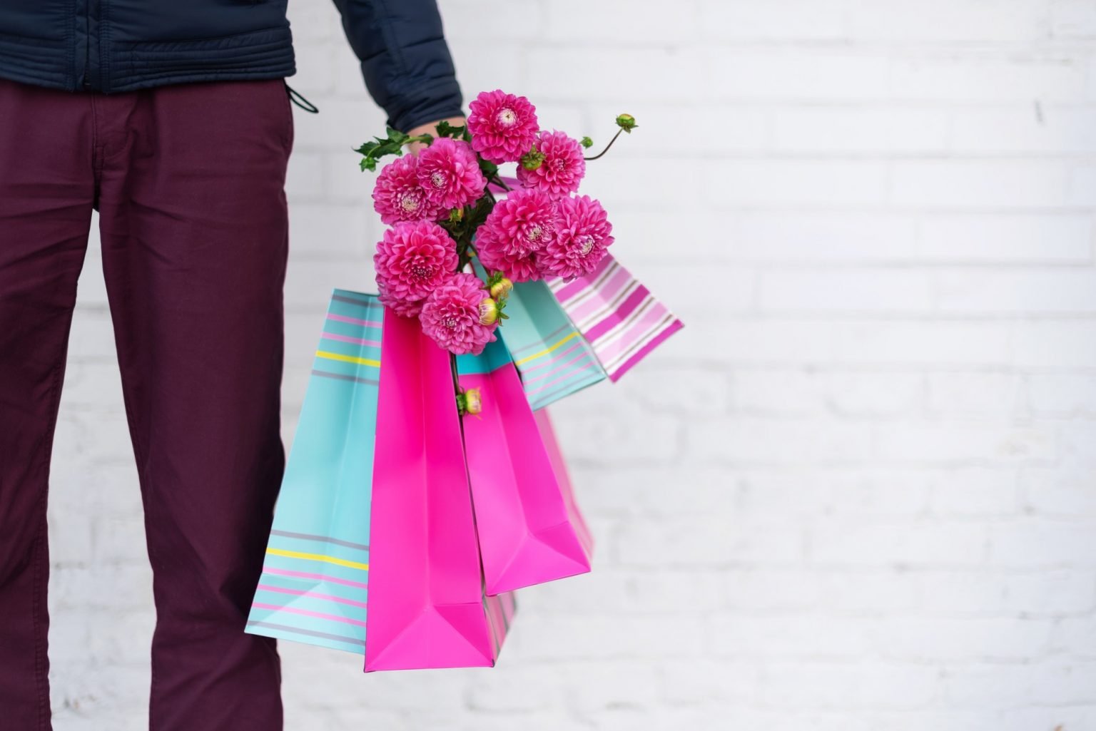 Stylish man holding flowers and pink, blue shopping bags on brick background. Woman's day