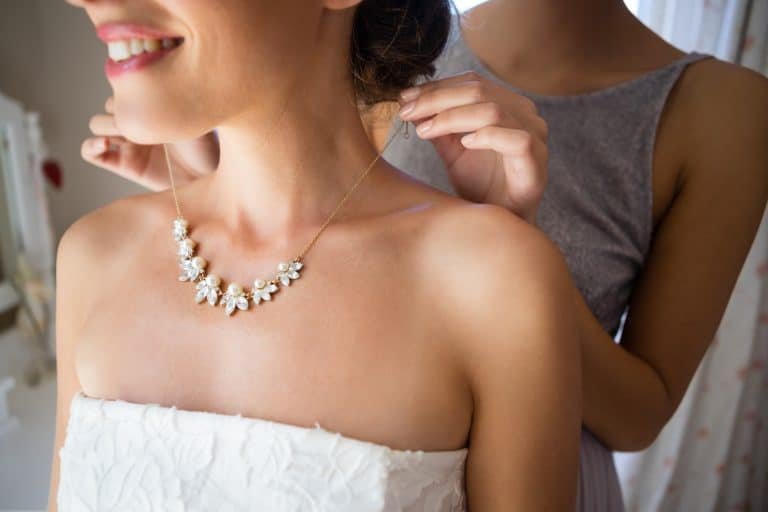 Midsection of bridesmaid fastening bride necklace in room